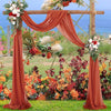A rustic wedding arch decorated with orange chiffon draping fabric panels and floral arrangements in a garden setting.