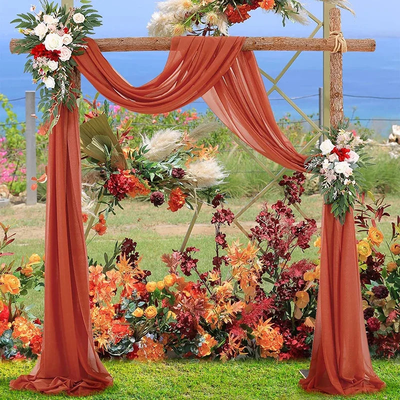 A rustic wedding arch decorated with orange chiffon draping fabric panels and floral arrangements in a garden setting.
