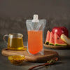 A clear liquid drink is contained in a resealable pouch on a wooden board with a glass jug and a slice of watermelon.