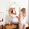 A woman is looking in a large round vanity mirror with a matte metal frame in a bright bathroom setting.
