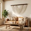A bohemian macrame wall tapestry hangs on a white wall above a natural leather sofa and wooden coffee table.