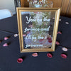 A framed glass vase filled with pink and white flowers sits on a black tablecloth surrounded by scattered rose petals.