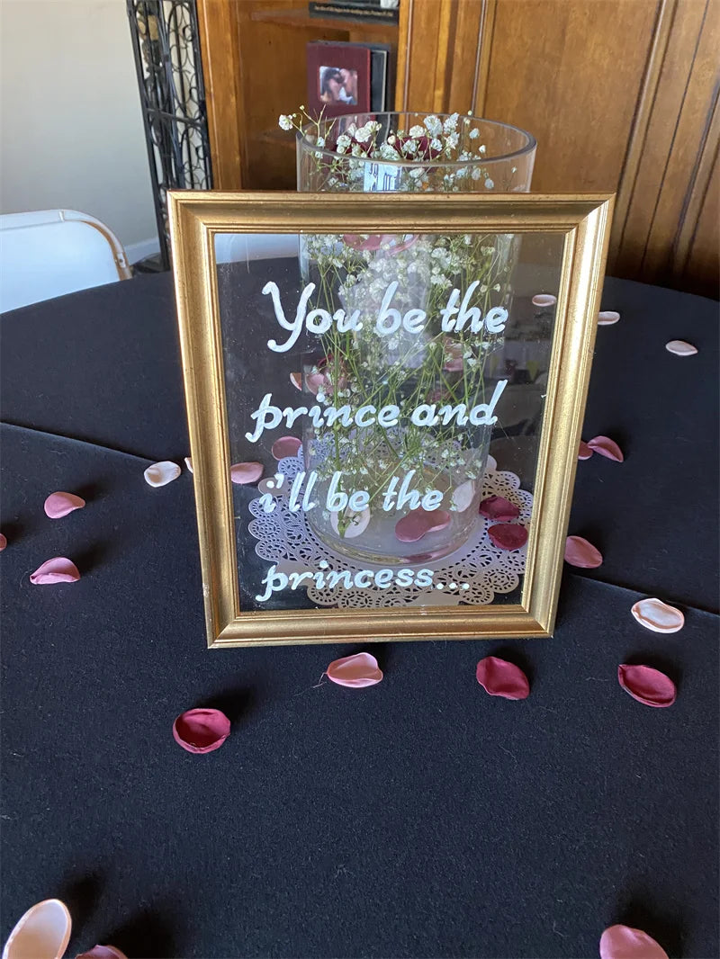 A framed glass vase filled with pink and white flowers sits on a black tablecloth surrounded by scattered rose petals.