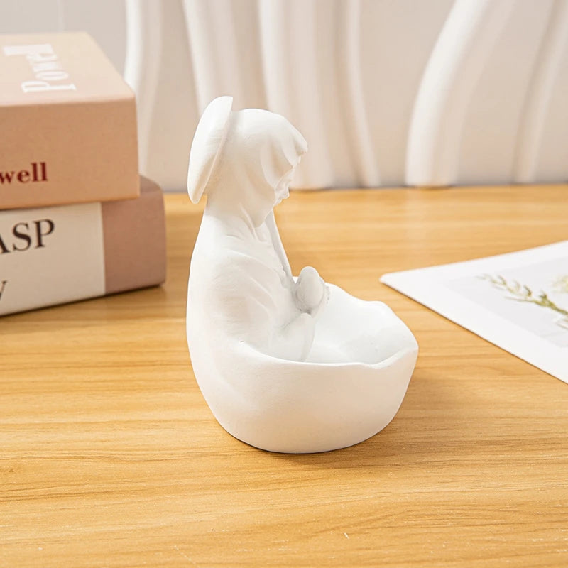 A white resin statue of the Virgin Mary in a seated pose, displayed on a wooden table.