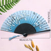 A light blue bamboo folding fan with white floral designs and a blue tassel displayed on a pink background.