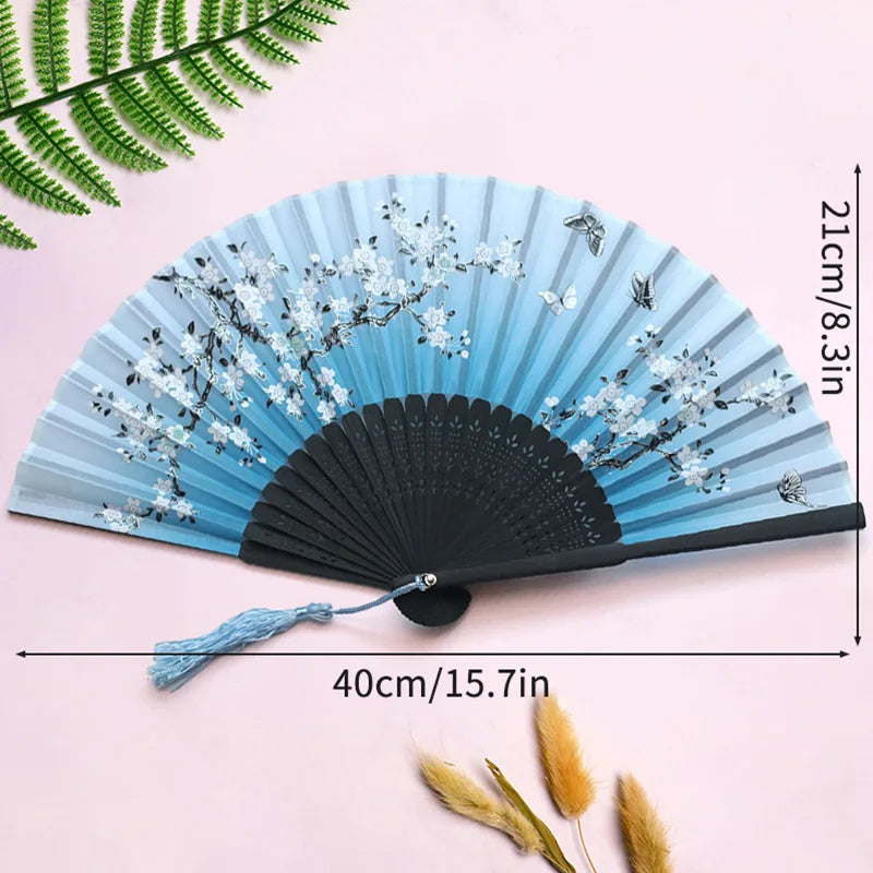 A light blue bamboo folding fan with white floral designs and a blue tassel displayed on a pink background.
