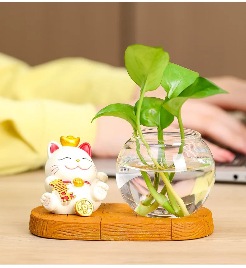 A clear glass vase holds a green ivy plant and a small white lucky cat figurine on a wooden base.