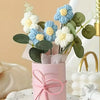 A handmade floral bouquet featuring light blue and cream flowers in a pink cylindrical vase on a white speckled plate.