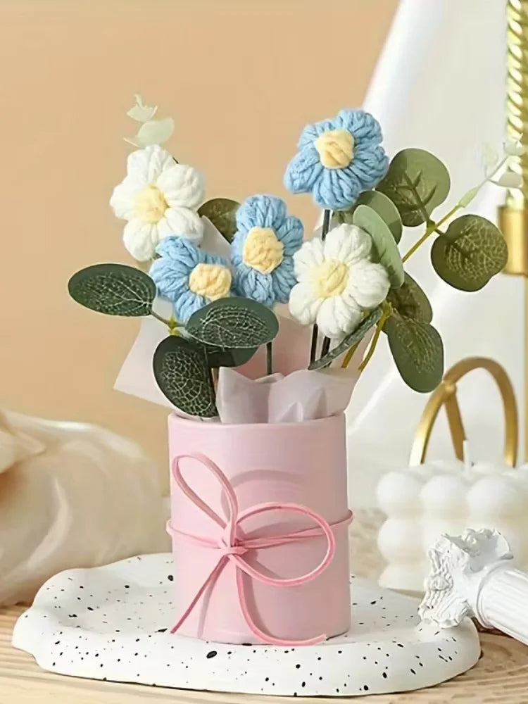 A handmade floral bouquet featuring light blue and cream flowers in a pink cylindrical vase on a white speckled plate.