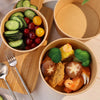 A wooden tray holds disposable kraft paper lunch bowls filled with various fresh vegetables and seafood.