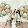 A woman in a light green dress sits surrounded by a large arrangement of sage green and gold confetti balloons.