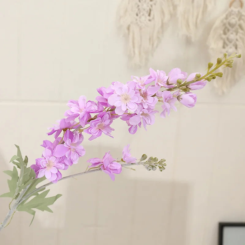 Une branche de fleurs de delphinium roses artificielles avec des feuilles vertes, présentées sur un fond clair.