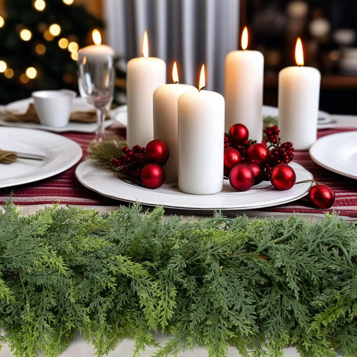 A festive holiday table setting featuring white candles, red ornaments, and a green cedar garland.