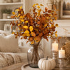 A bouquet of artificial fall eucalyptus stems with warm orange and yellow leaves displayed in a glass vase on a wooden surfac