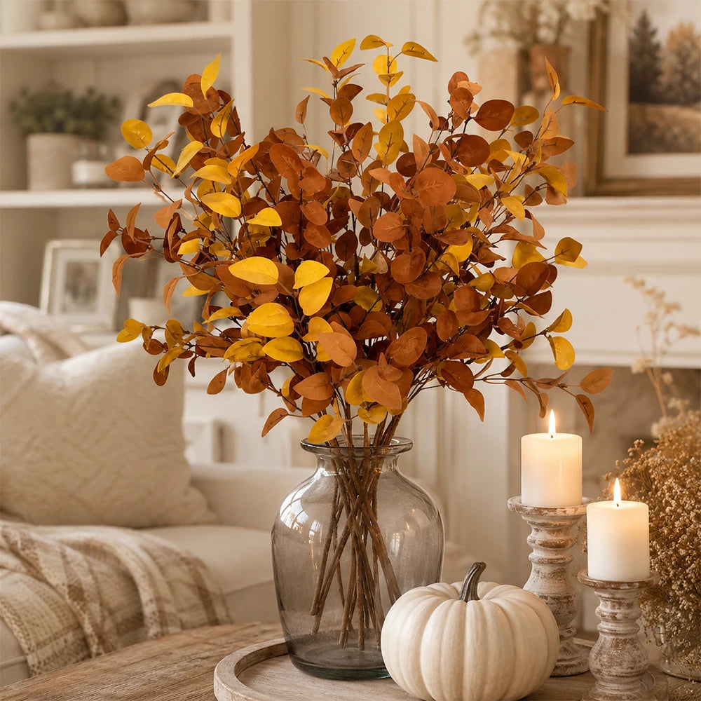 A bouquet of artificial fall eucalyptus stems with warm orange and yellow leaves displayed in a glass vase on a wooden surfac