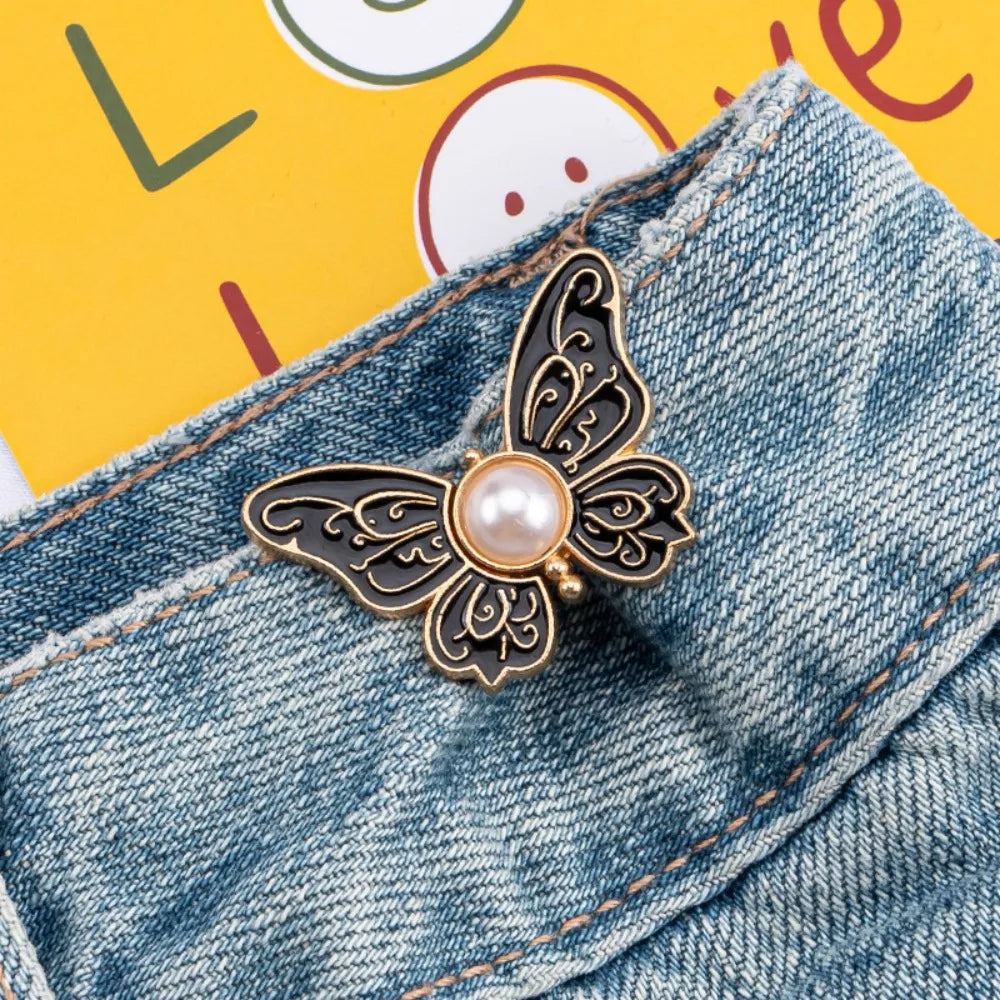A gold-toned butterfly clip with a pearl accent is fastened onto the denim fabric of pants.
