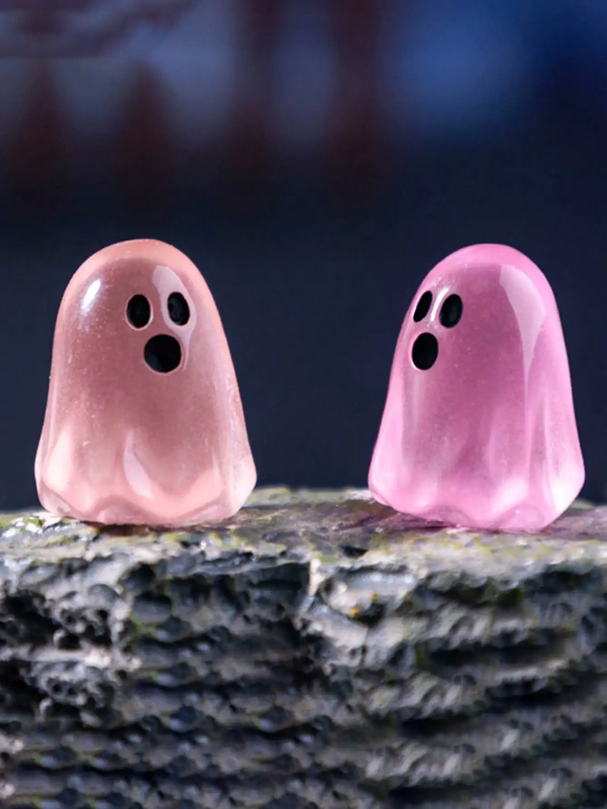 Two translucent pink ghost figurines resting on a rough gray surface against a dark blue background.