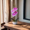 A realistic artificial orchid potted plant with vibrant purple flowers sits on a wooden table.