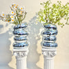 A decorative vase holds white daisies and green foliage, featuring a stacked silver mirror tile design.