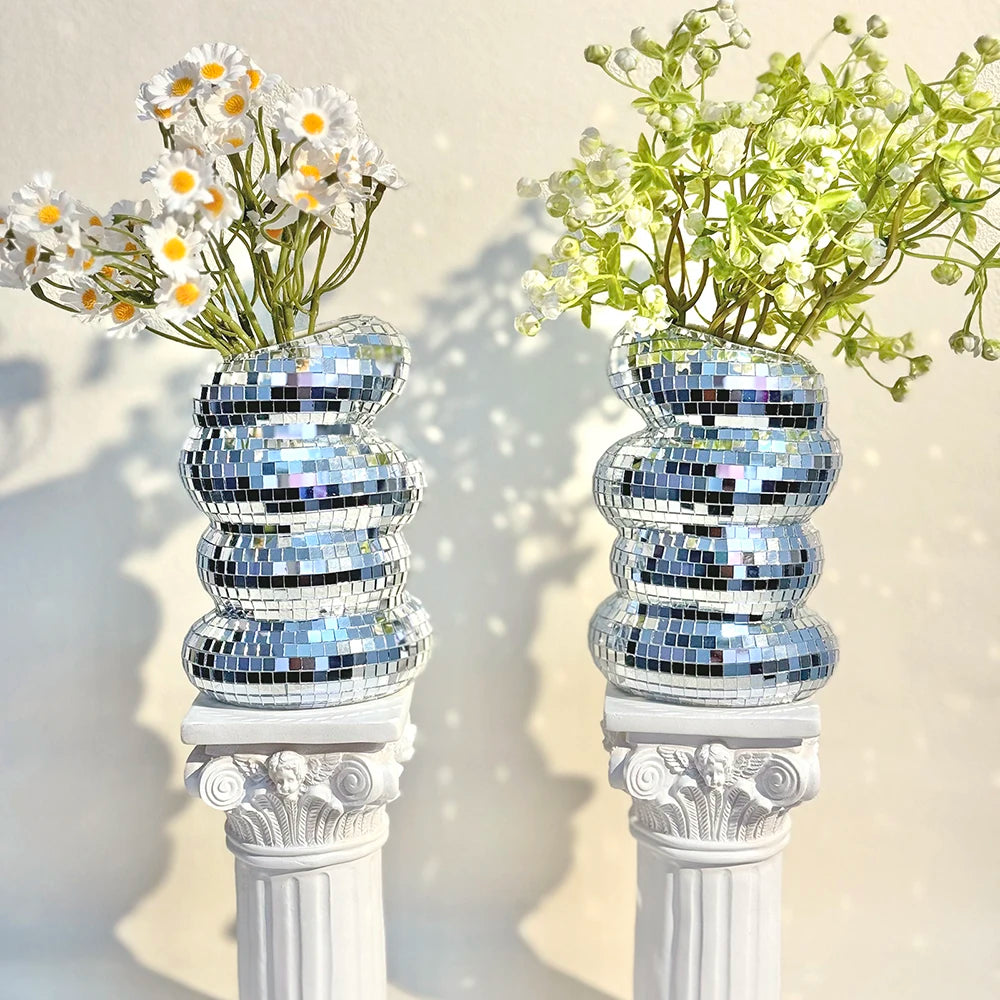 A decorative vase holds white daisies and green foliage, featuring a stacked silver mirror tile design.