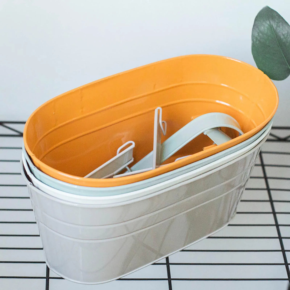 A modern boho metal hanging planter with an orange top and a light gray body sits on a black wire grid.