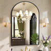 An arched wall-mounted bathroom mirror with an aluminum frame reflecting a chandelier and window.