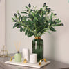 A vase filled with artificial olive leaf branches on a light gray table.
