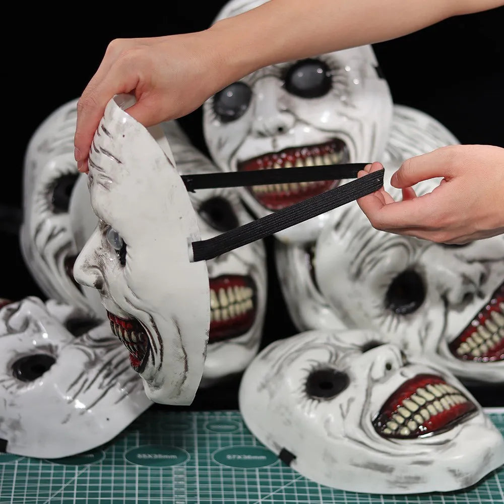 A hand is holding a white porcelain demon mask with painted features, suggesting a Halloween cosplay prop.