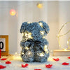 Flauschiger künstlicher Rosen-Teddybär mit warmen Lichterketten auf weißem Untergrund.