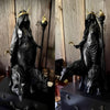 A dark resin statue of a goddess with a crown and staff, standing over a black animal.
