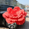 A vibrant red artificial peony flower is displayed in front of a dark car.