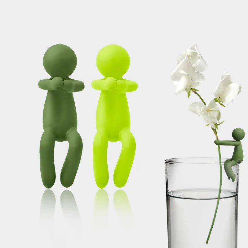 A set of three human-shaped plant supports in shades of green and lime yellow, displayed next to a glass of water.