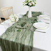 A sage green semi-sheer gauze table runner draped on a white tablecloth with white plates and silverware.