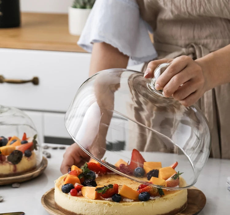 A glass dome cover is placed over a cake tray, showing a round cake decorated with fresh fruit.