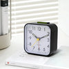 A black digital alarm clock with a white face and green accents sits on a white surface.