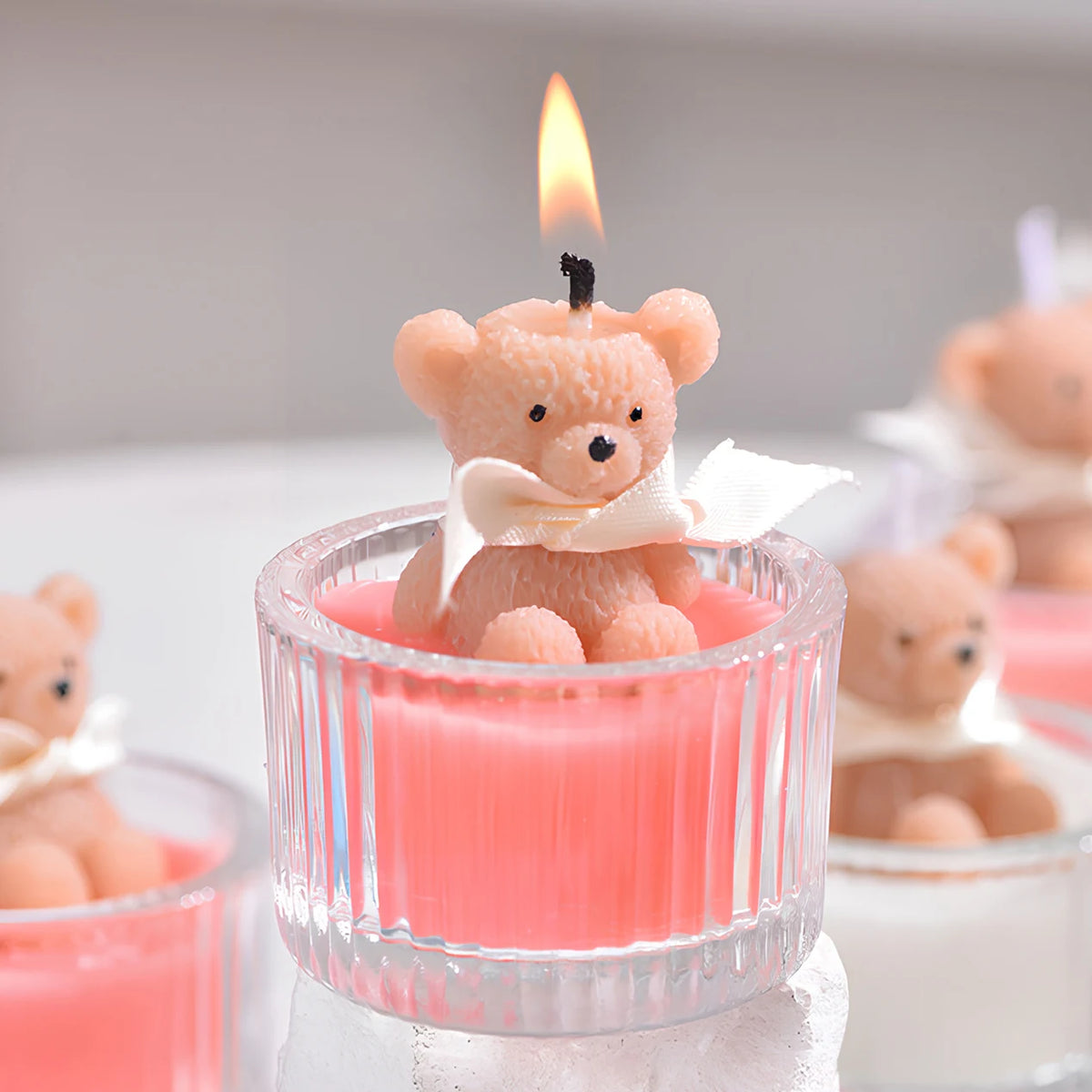A small teddy bear candle in a glass jar with a ribbon is lit, set against a white background.
