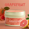 A jar of grapefruit scented soy wax travel candle with fresh grapefruit fruit nearby.
