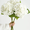 A hand holds a bouquet of white hydrangea silk flower branches with green leaves.