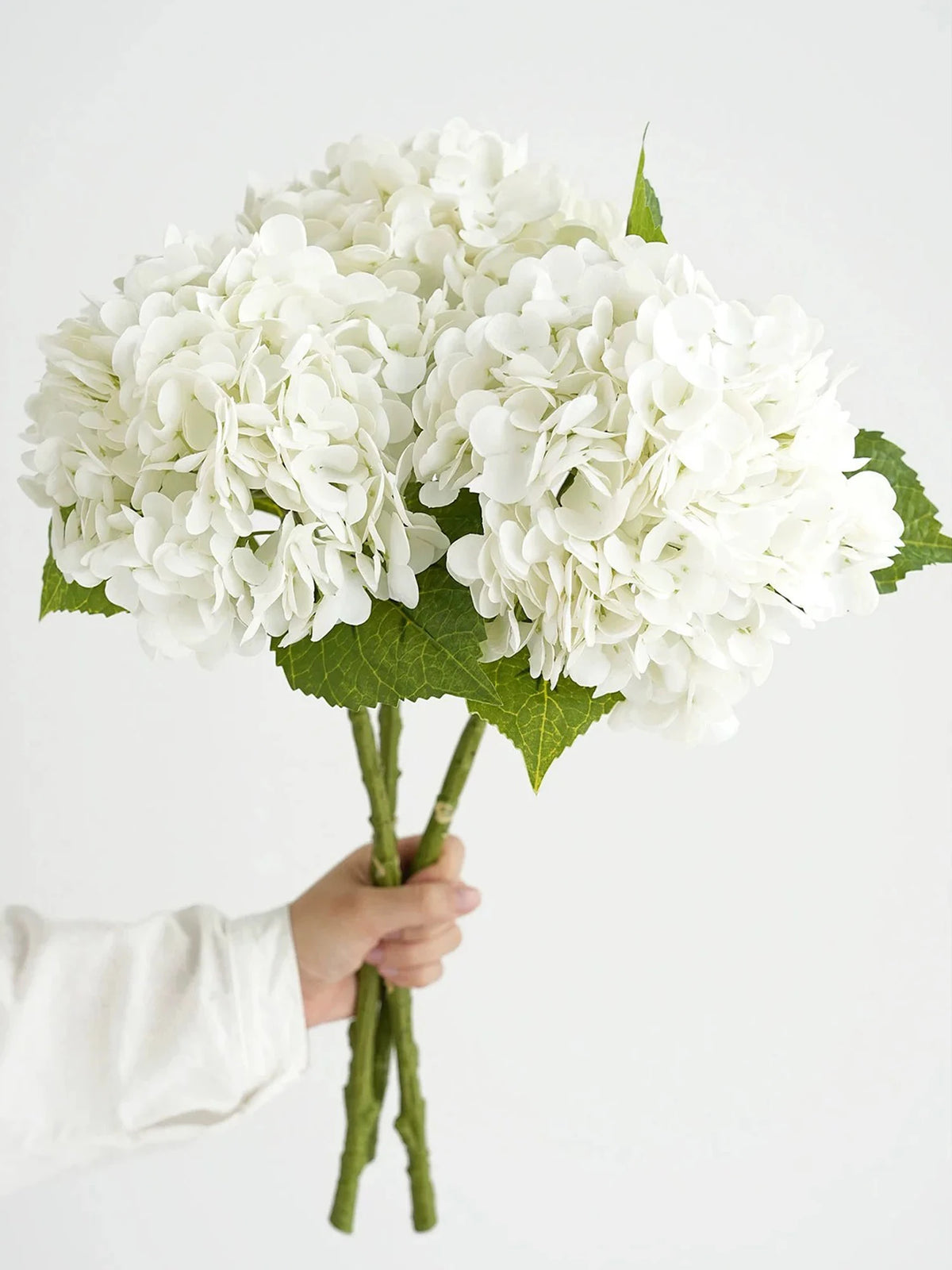 A hand holds a bouquet of white hydrangea silk flower branches with green leaves.