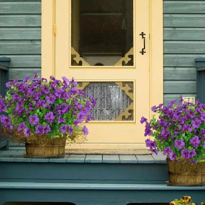 A close-up of purple petunias in terracotta pots on a porch ledge next to a light yellow door.