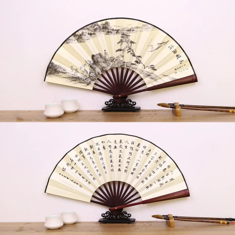 A vintage folding fan with a black and cream design featuring a detailed landscape, displayed on a wooden surface.