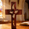 A wooden cross carved with intertwined hearts resting on a wooden surface in a church setting.