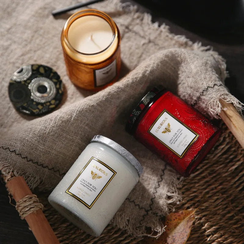 A hand-poured natural soy candle in a glass jar sits on a rustic linen cloth next to a red jar and a small decorative item.