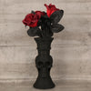 A black skull-shaped decorative vase holds a bouquet of red roses and dark green leaves.