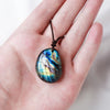 A hand holds a natural labradorite pendant necklace with iridescent blue and green colors.