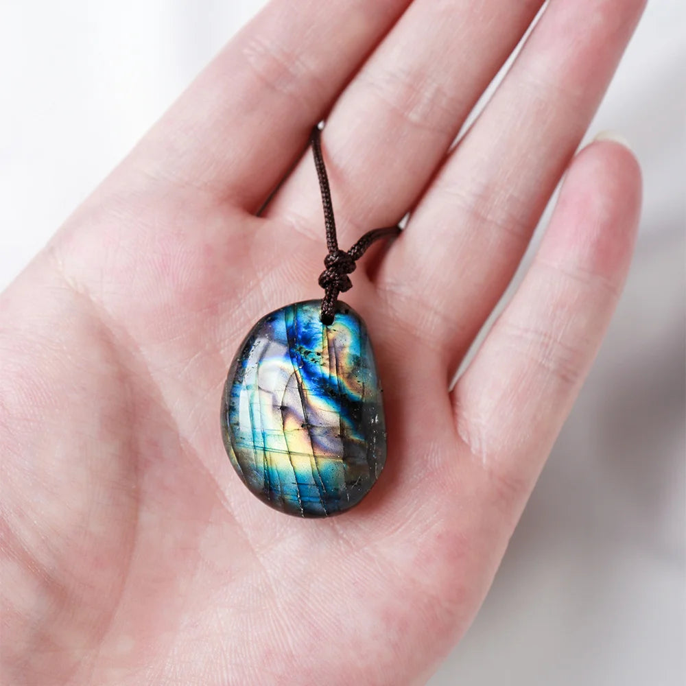 A hand holds a natural labradorite pendant necklace with iridescent blue and green colors.