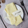 A set of ten yellow handcrafted fringed cotton napkins is displayed on a white plate with a tag that reads Je t'aime