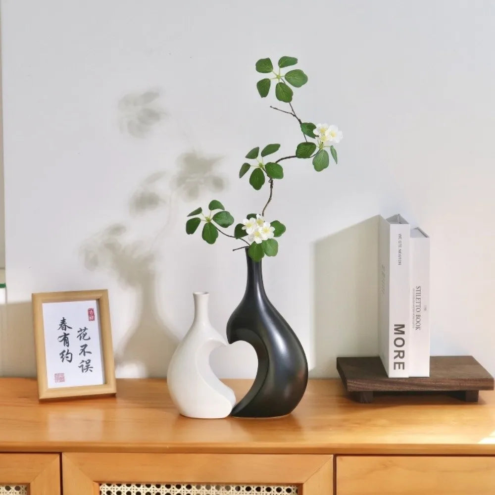 A black heart-shaped ceramic vase holds a branch of green leaves and white flowers on a wooden table.