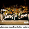 A glowing neon sign spelling Cheers to Love is displayed on a dark background with floral accents.