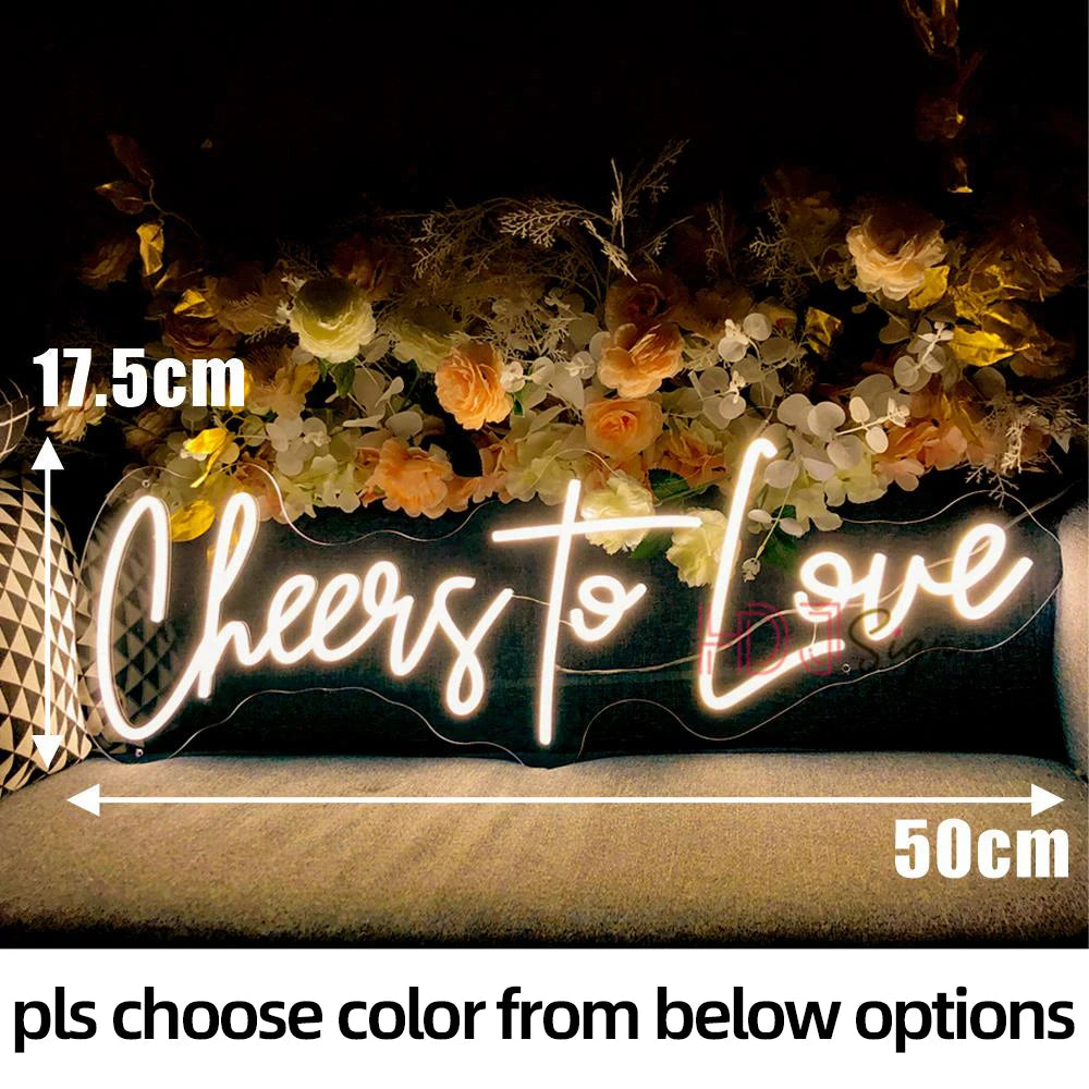 A glowing neon sign spelling Cheers to Love is displayed on a dark background with floral accents.